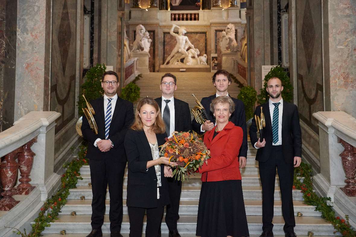 Katrin Völk überreicht Sabine Haag einen Blumenstrauß auf der Foyerstiege des KHM, hinter ihnen stehen Bläser.