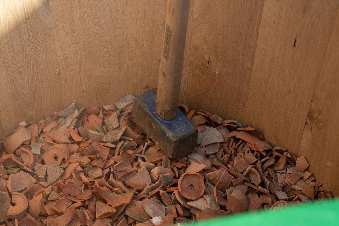 Mit einem Hammer werden Tonstücke in einem Kübel zu kleinen Stücken zerstoßen