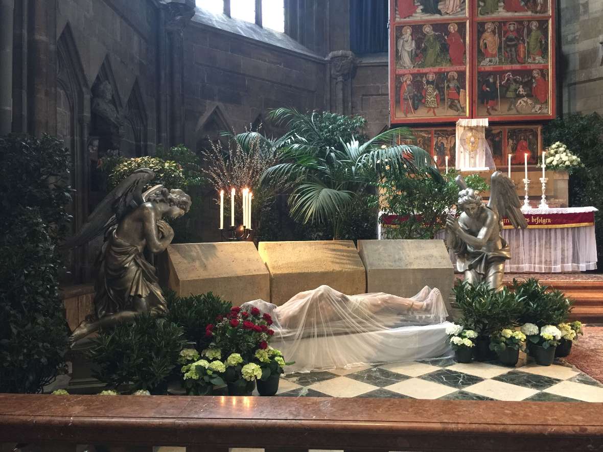 zu sehen ist der Innenraum einer Kirche mit einem Altar, geschmückt mit weißen Blumen und Grünpflanzen; vor dem Altar sieht man eine Statue, die verhüllt mit einem weißen Stoff liegt;