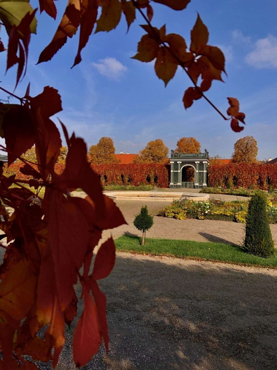 zu sehen ist ein herbstlicher Park, Bäume mit rot gefärbten Laub und leere Erdbeete