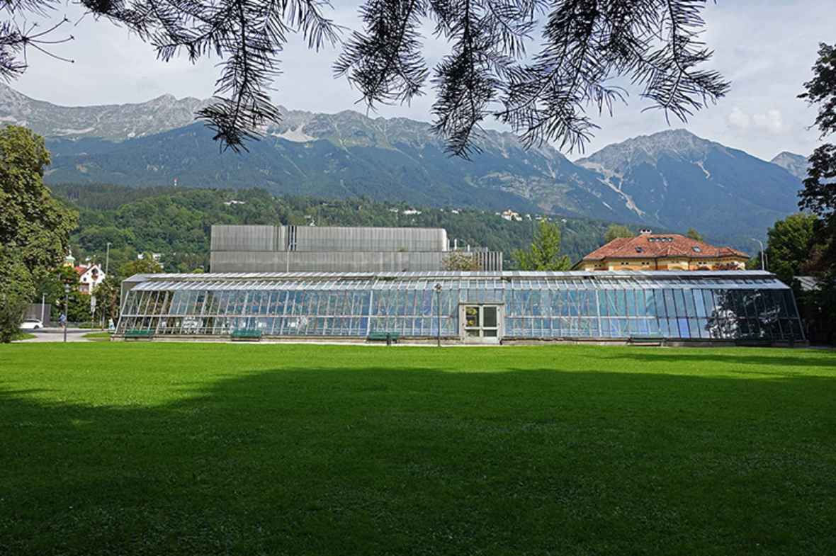 zu sehen ist ein großes Glashaus; im Vordergrund eine grüne Wiese; im Hintergrund Berge